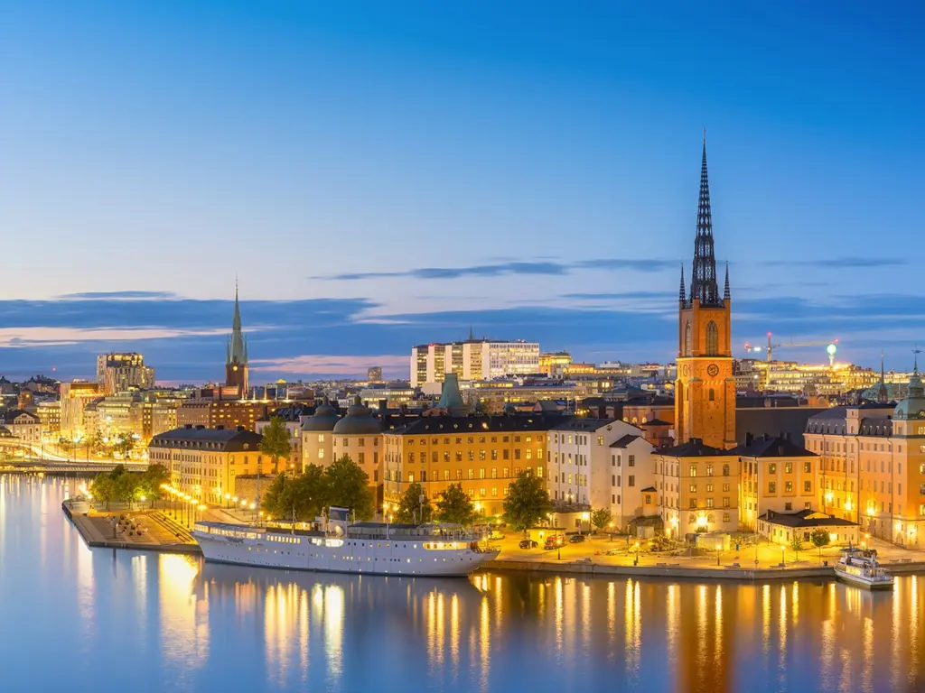 Panoramavy over centrala Stockholm vid vatten i kvällsbelysning med Riddarholmskyrkans torn och upplysta byggnader – symbol for lokal IT‑partner med stabil drift och lang erfarenhet.