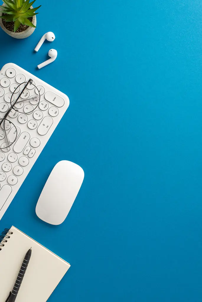 Skrivbord sett ovanifrån med tangentbord, mus, hörlurar, anteckningsblock och växt.