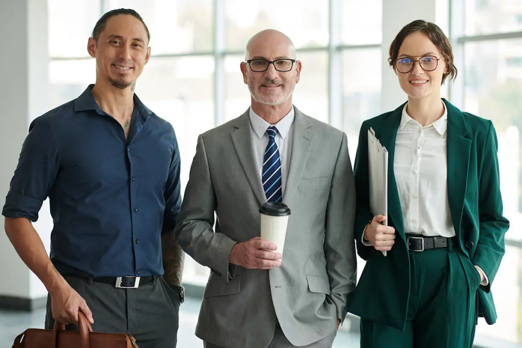Tre personer i affärskläder står i en ljus kontorsmiljö med dokument och kaffemugg