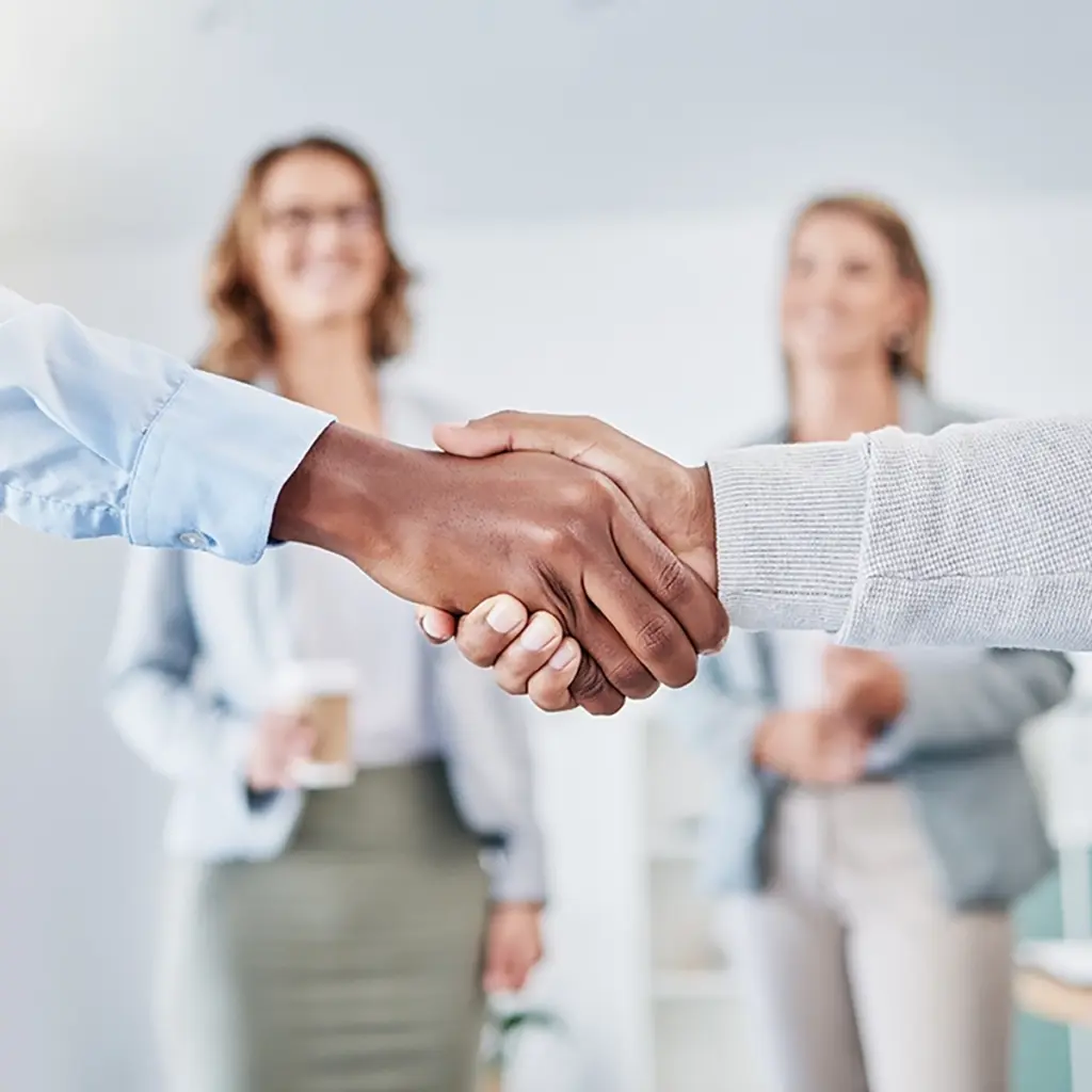Två personer skakar hand i förgrunden medan två personer står suddigt i bakgrunden i en kontorsmiljö.