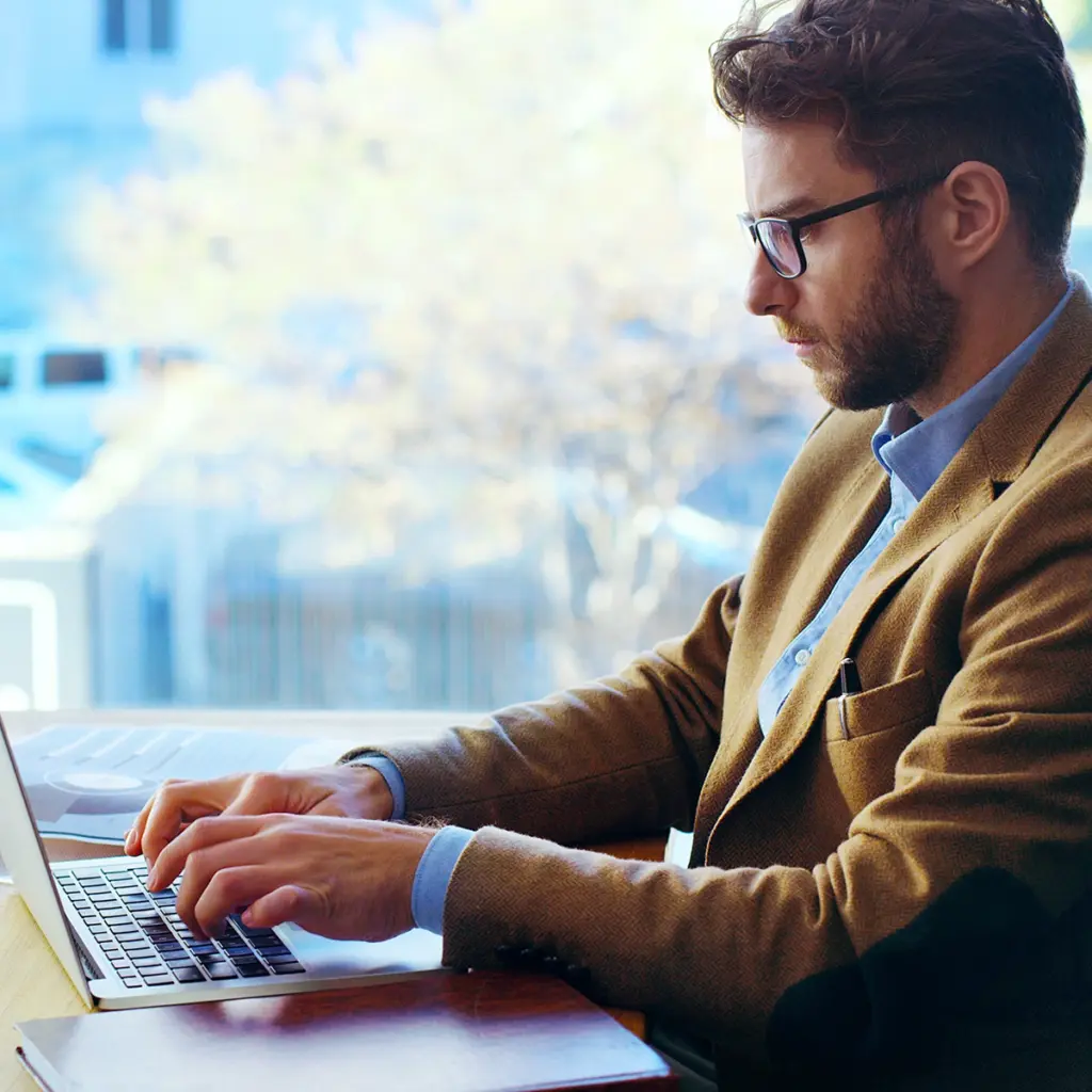 Person arbetar på laptop vid bord nära stort fönster.