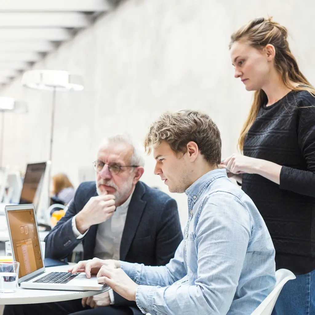 Tre personer samlas runt en laptop i ljus kontorsmiljö.
